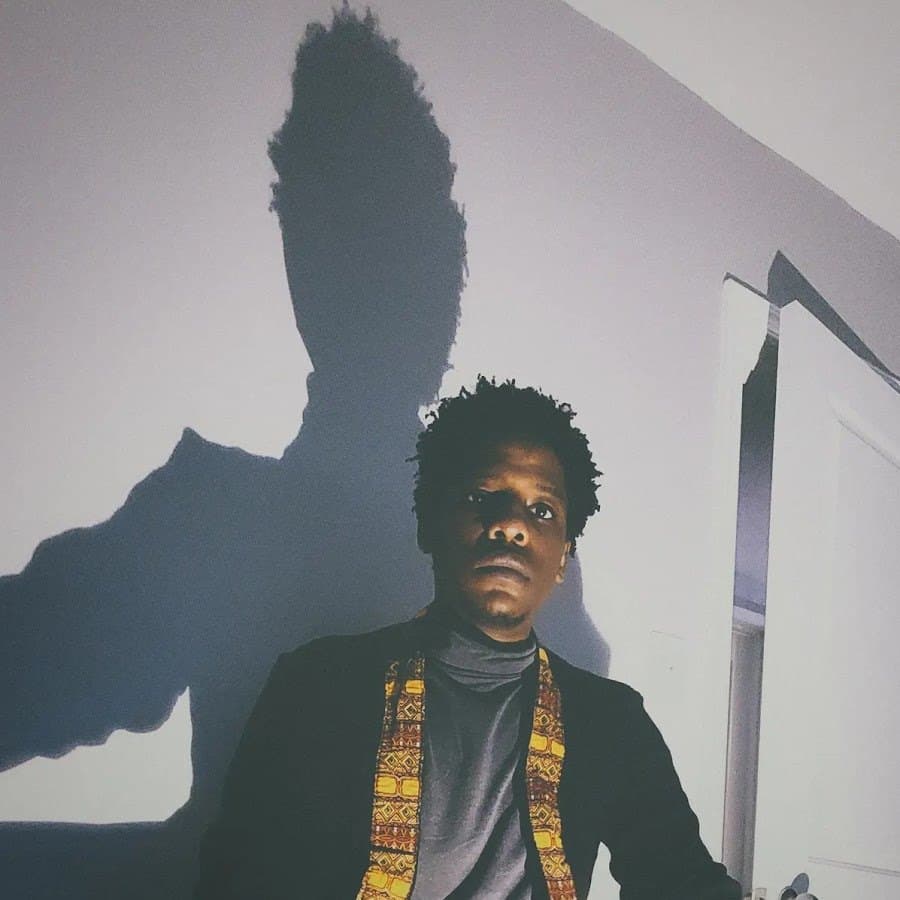 Simbai against a pale wall with his shadow behind him, wearing a patterned yellow scarf