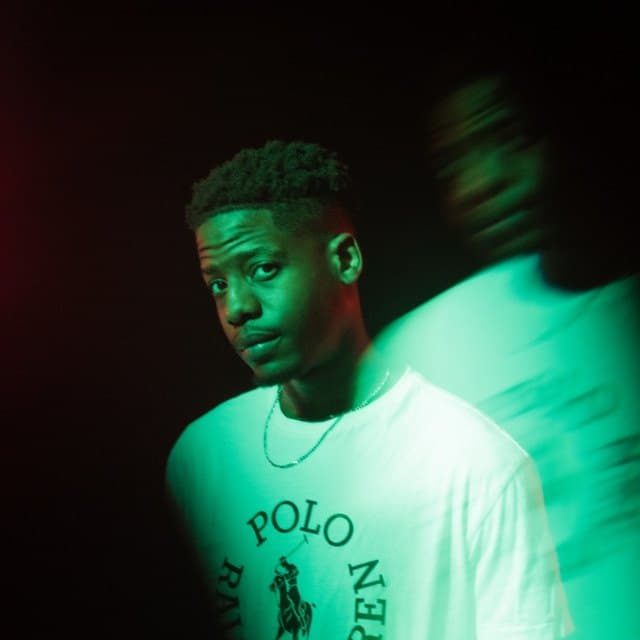 Simbai in a white T-shirt under green light, a blurred figure moving past him