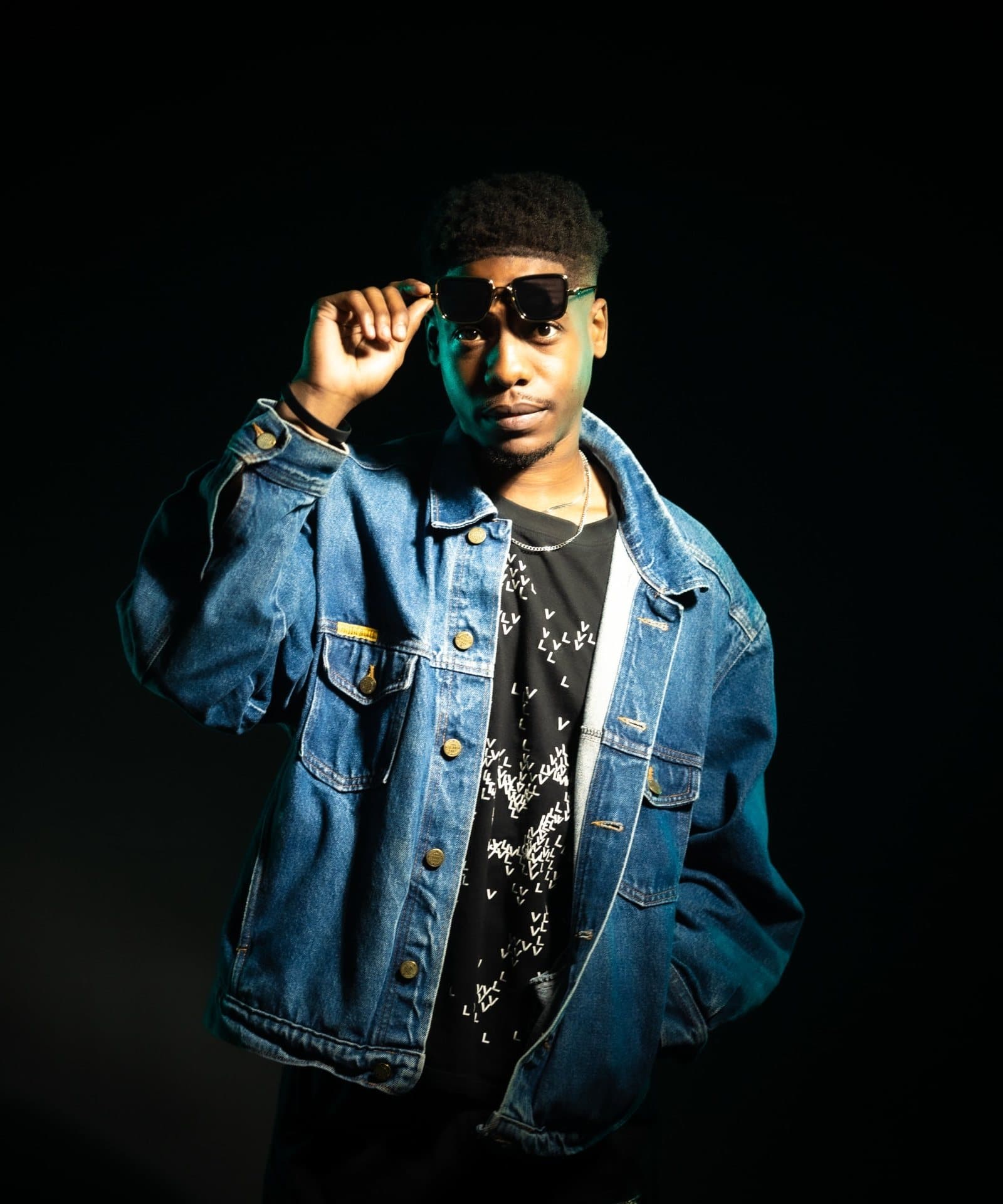 Simbai in a denim jacket, lifting his sunglasses, lit in cool blue on black